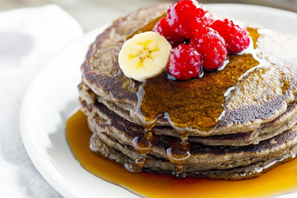 Stack of buckwheat pancakes with banana and raspberries, covered in maple syrup, served on a white plate.