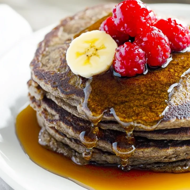 Stack of buckwheat pancakes with banana and raspberries, covered in maple syrup, served on a white plate.