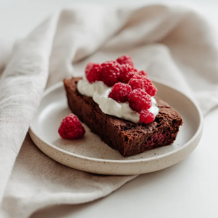 Moist chocolate loaf slice topped with fresh raspberries and yogurt on a speckled plate