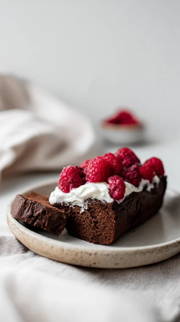 A close-up slice of chocolate raspberry loaf topped with yogurt and fresh raspberries on a ceramic plate
