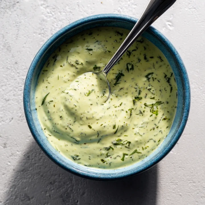 Top-down view of lemon herb tahini sauce in a blue-rimmed bowl with spoon, creamy texture and fresh herb garnish