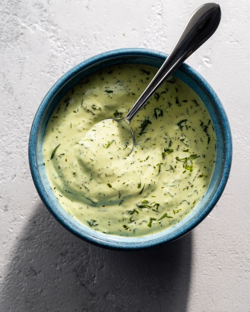 Top-down view of lemon herb tahini sauce in a blue-rimmed bowl with spoon, creamy texture and fresh herb garnish